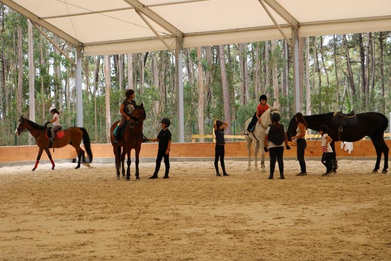 Jovens a montar a cavalo durante a sessão prática no picadeiro do campo de férias de equitação