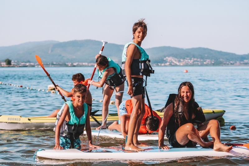  Na imagem, um grupo de jovens está a divertir-se em pranchas de paddle e caiaques no lago, todos com coletes salva-vidas e sorrisos. Esta atividade aquática no campo de férias Serra da Estrela oferece momentos de pura alegria e aventura na água.