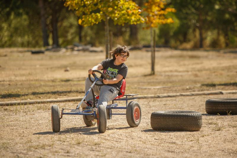  Na imagem, uma jovem está a conduzir um kart a pedais ao ar livre, contornando obstáculos feitos de pneus no chão. Esta atividade no campo de férias monsanto oferece diversão e adrenalina num cenário natural.