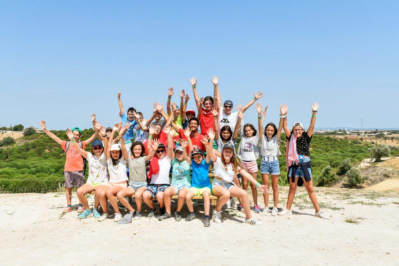grupo de participantes reunidos no campo de férias a sorrir e com os braços levantados