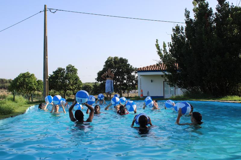  Na imagem, um grupo de jovens está numa piscina, cada um segurando uma bola de praia azul e branca. Ao fundo, um monitor observa, enquanto os participantes parecem estar a divertir-se numa atividade aquática. Esta cena vibrante e animada reflete o ambiente descontraído e interativo do curso de inglês em Braga, onde aprender é uma experiência refrescante e cheia de energia.