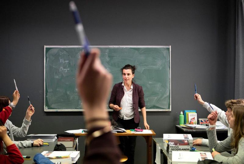  uma professora em frente a um quadro preto numa sala de aula, com estudantes que levantam as mãos a segurar canetas, como se estivessem prontos a participar. Este ambiente dinâmico reflete a interação e entusiasmo que encontrarás no curso de línguas em Coimbra.