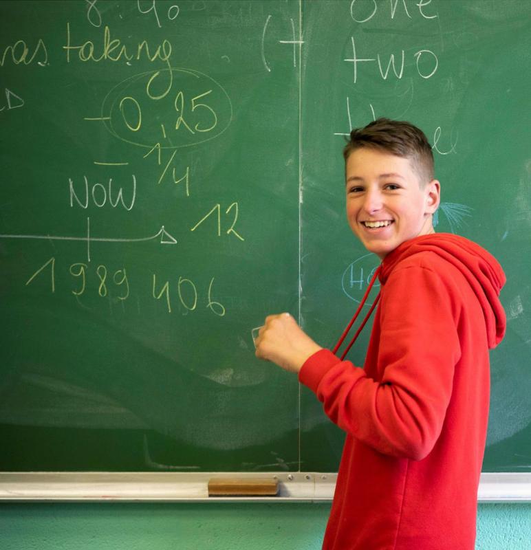  Na imagem, um jovem com um sorriso confiante está junto a um quadro verde com várias anotações escritas a giz. O ambiente é de participação ativa e alegria, transmitindo a energia e motivação do curso de línguas em Coimbra.