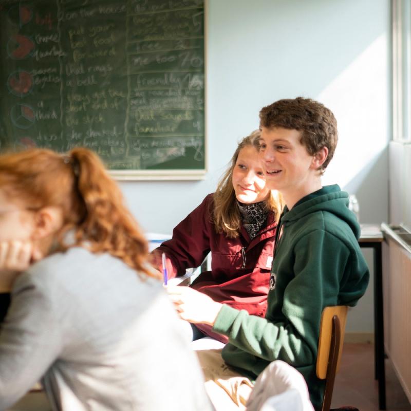  Na imagem, três jovens estão numa sala de aula iluminada pela luz do sol, com expressões descontraídas e sorridentes, enquanto participam de uma atividade. Ao fundo, há um quadro verde com várias frases escritas, transmitindo o ambiente acolhedor e colaborativo do curso de línguas em Coimbra.