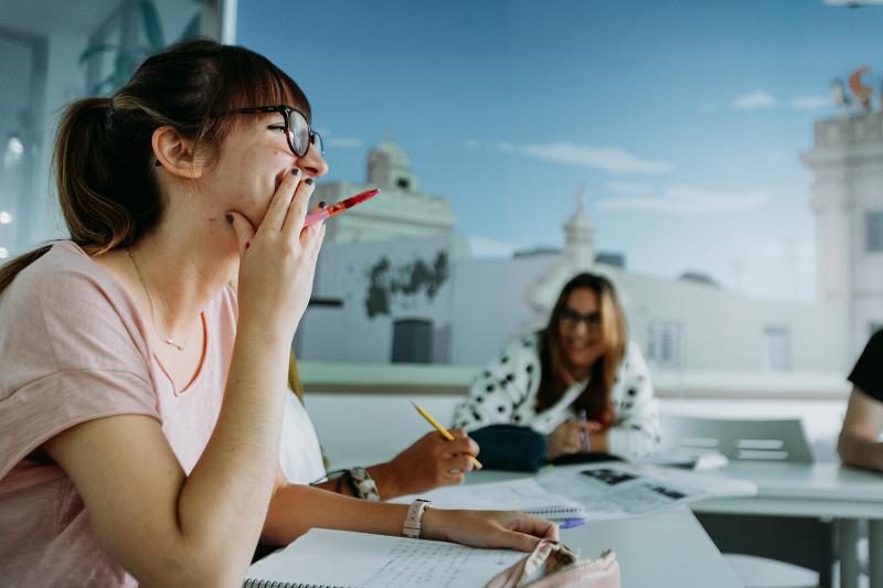 Aluna a sorrir numa aula envolvente dos cursos de língua espanhola, com colegas ao fundo.