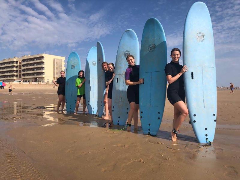 Alunas dos cursos de espanhol a sorrir na praia, segurando pranchas de surf alinhadas.