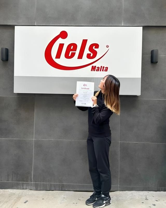 Participante a segurar o certificado à frente do logo, celebrando a conclusão dos cursos de inglês em malta.