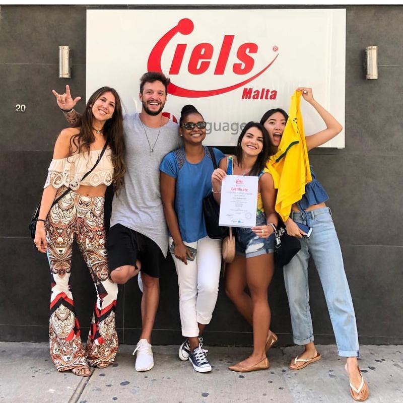 Participantes a posar na parede à frente do logo, celebrando a experiência dos cursos de inglês em malta.