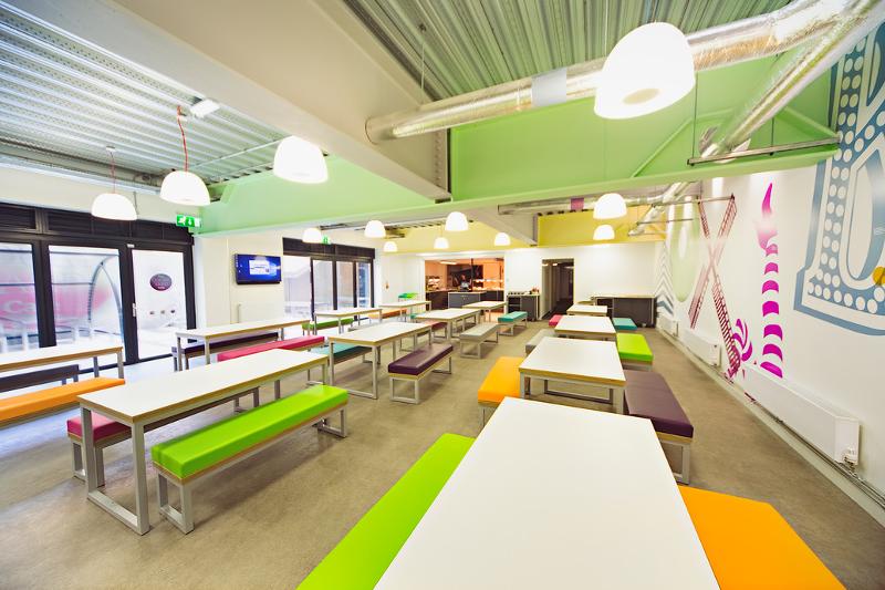 Um refeitório vibrante e moderno, equipado com mesas e bancos coloridos, ideal para socializar enquanto desfruta de refeições durante o curso de ingles para jovens.