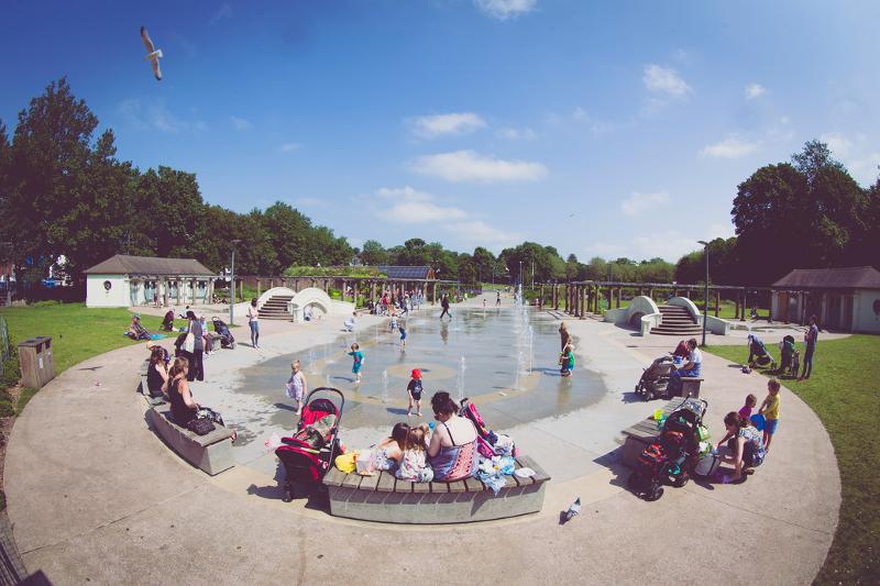 Crianças e famílias desfrutam de um dia ensolarado num parque com fonte interativa, ideal para jovens que querem praticar ingles num ambiente divertido.