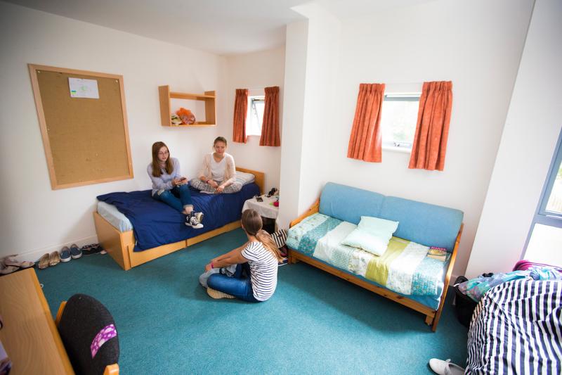 Três jovens conversam num quarto confortável com camas e sofá, parte de um alojamento para curso de ingles para jovens.