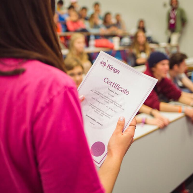 Aluna segura um certificado enquanto outros estudantes assistem na sala, celebrando a conclusão de um curso de língua inglesa.