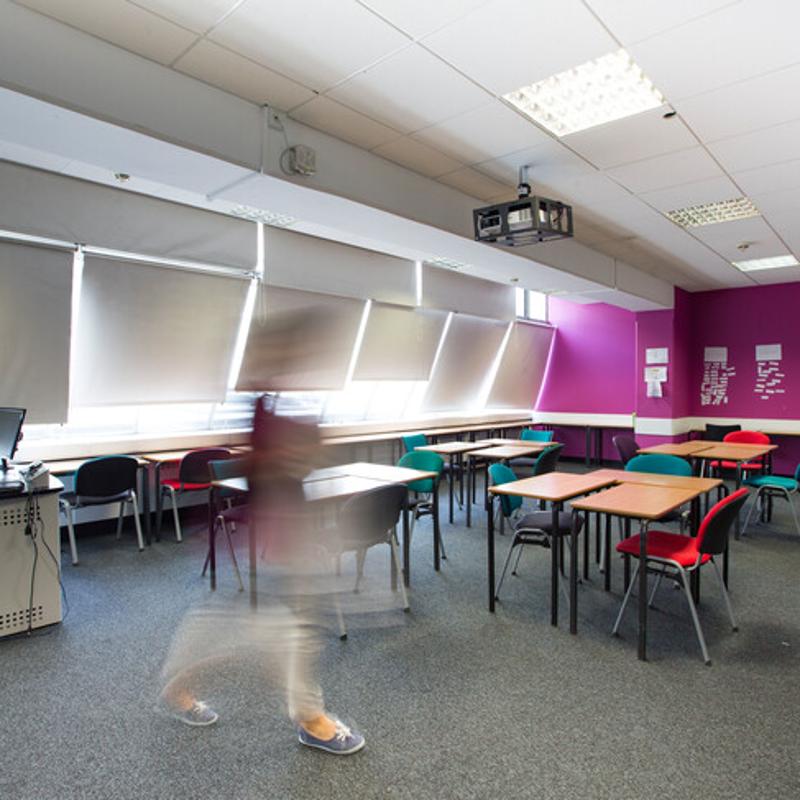 Sala de aula com mesas e cadeiras coloridas, onde uma aluna se move rapidamente, indicando um ambiente dinâmico de aprendizagem no curso de língua inglesa.
