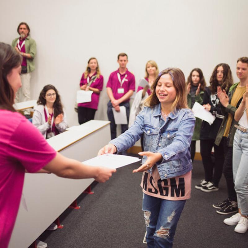 Estudante sorridente a receber um certificado de participação no curso de inglês em londres 4 semanas, enquanto colegas aplaudem.