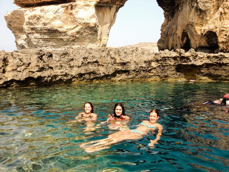 Três jovens a nadar em águas cristalinas junto a formações rochosas durante um curso de linguas em malta, desfrutando do ambiente natural.