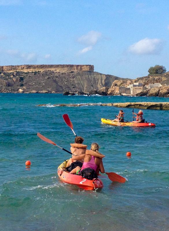 Jovens a remar em caiaques no mar como parte de atividades de um curso de linguas em malta, com paisagem costeira ao fundo.