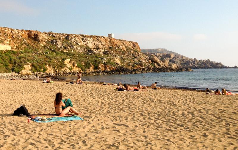  Pessoas relaxam numa praia de areia dourada com colinas rochosas ao fundo durante um curso de linguas em malta.