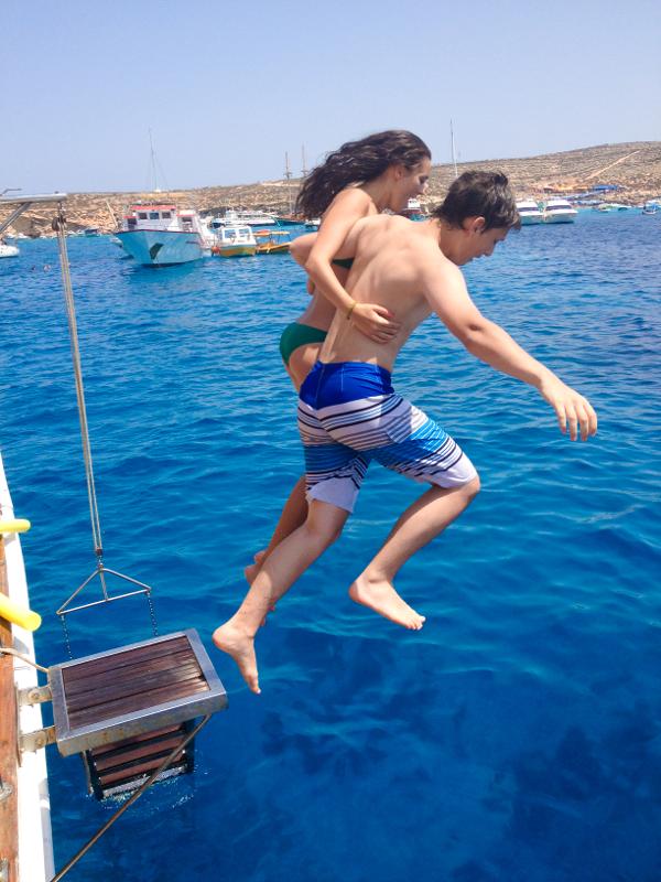Dois jovens a saltar juntos de um barco para águas azuis cristalinas durante um curso de linguas em malta.
