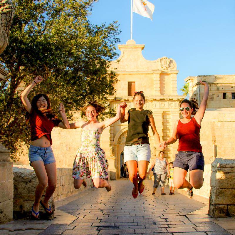 Quatro jovens a saltar de alegria em frente a um monumento histórico em malta durante um curso de linguas.