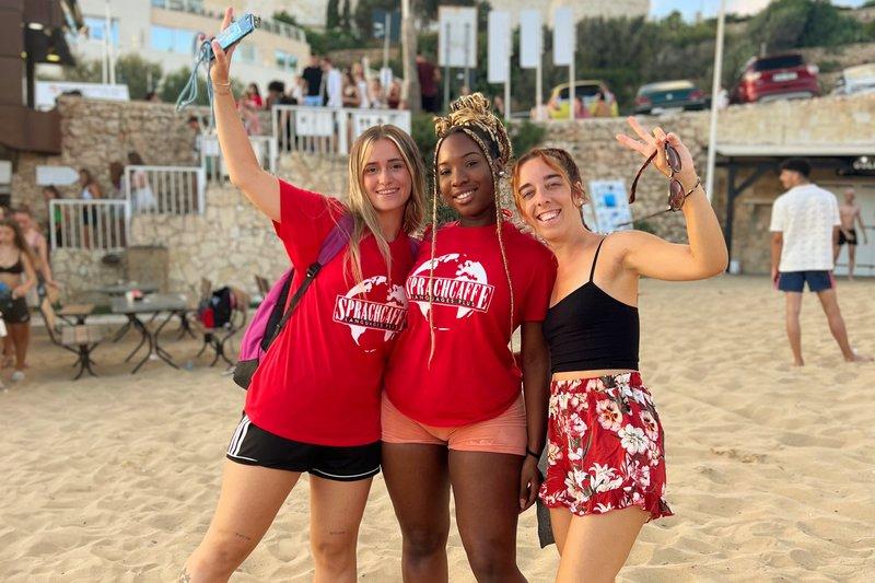 Três jovens sorrindo e a posar para a foto na praia, todas vestindo t-shirts vermelhas com o logo "Spracheffe", símbolo de uma escola de linguas, durante atividades de lazer num curso de ingles na Alemanha.