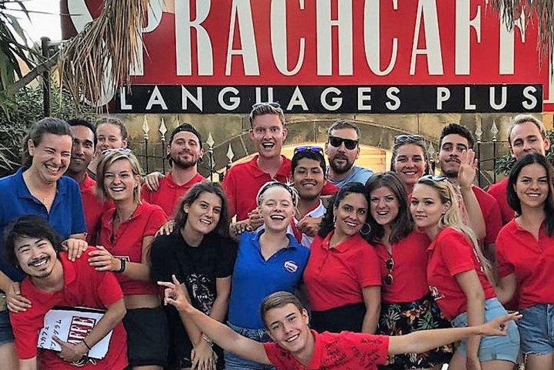 Grupo de estudantes e professores a sorrir juntos, mostrando a energia positiva do curso de linguas na alemanha. Todos estão a usar camisas vermelhas e há uma atmosfera descontraída.