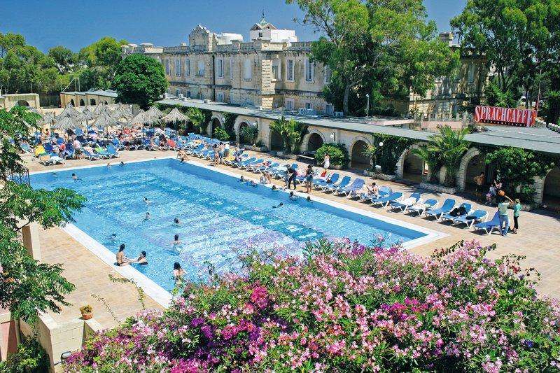 Vista de uma piscina rodeada por espreguiçadeiras e flores coloridas, com o edifício da escola de linguas ao fundo, criando um ambiente relaxante para o teu curso de linguas na alemanha.