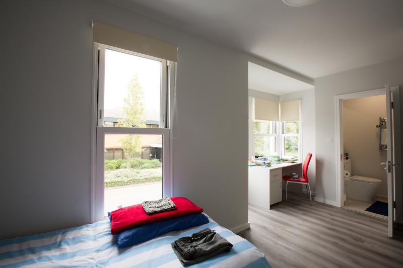 Quarto individual com cama, área de estudo e casa de banho privativa, parte do curso de ingles cambridge, oferecendo conforto e privacidade.