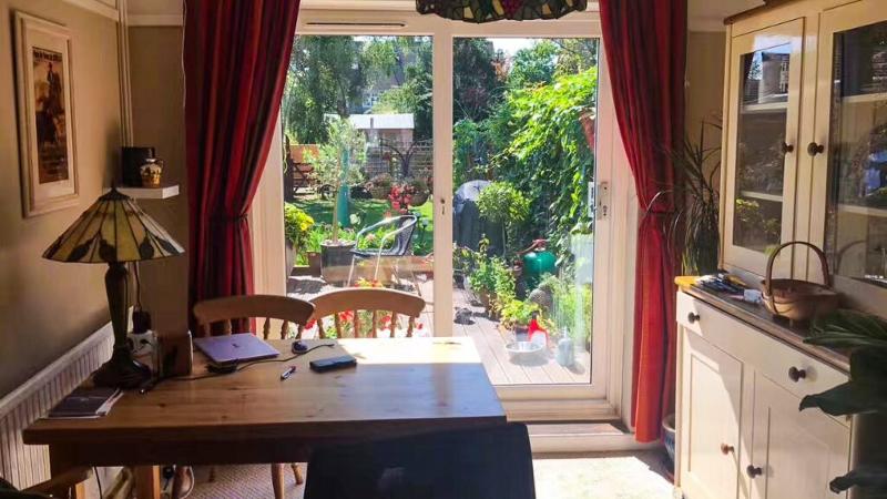 Sala iluminada com uma mesa de madeira e vista para um jardim florido, parte de uma casa que recebe participantes do curso de língua inglesa, proporcionando um ambiente acolhedor.