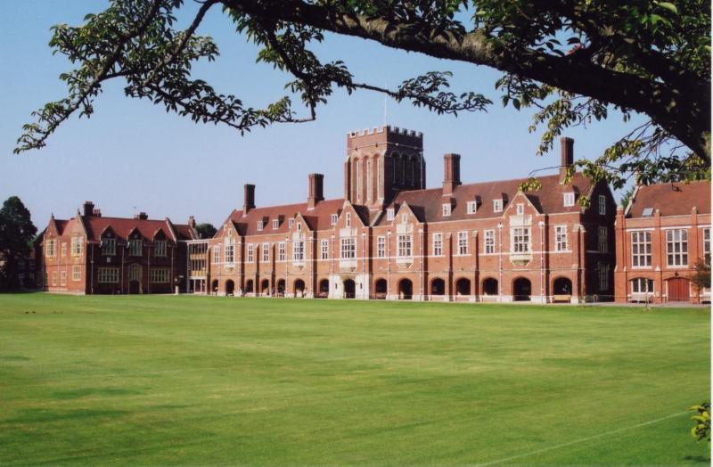Edifício histórico com torre e fachada de tijolos vermelhos, rodeado por um extenso campo verde, cenário ideal para um curso de língua inglesa.