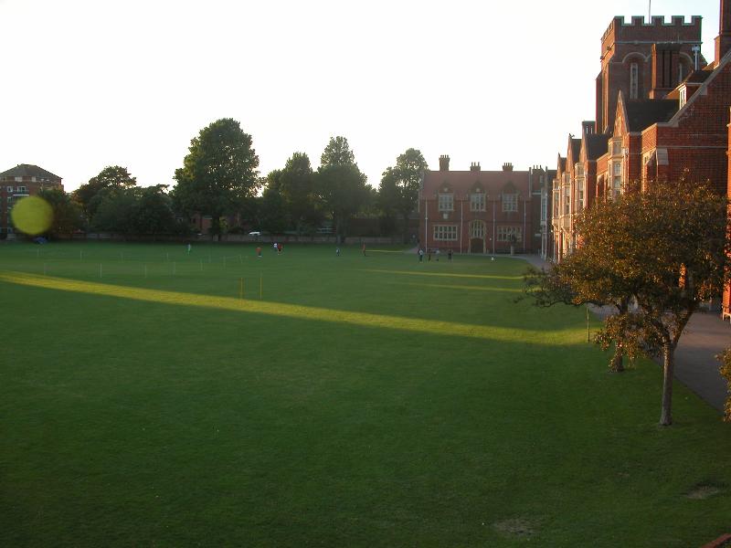 Campo verde ao pôr do sol num campus no Reino Unido, com edifícios de tijolos ao lado e algumas pessoas ao fundo.