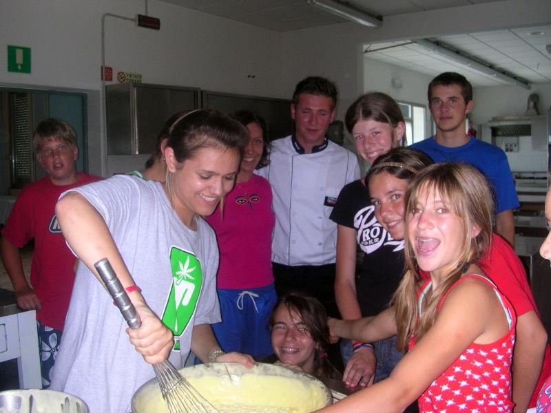 Grupo de jovens a aprender a cozinhar numa cozinha, com um chef e a preparar uma receita, enquanto participam num curso italiano.