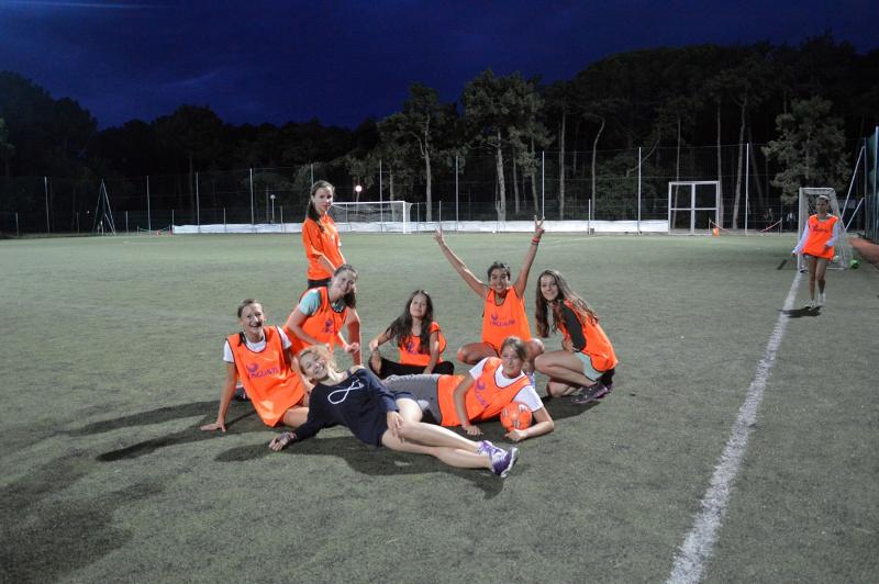 Grupo de jovens a divertir-se num campo de futebol, usando coletes e praticando desporto enquanto participam num curso italiano.