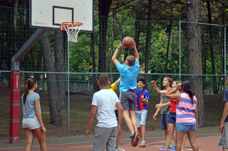 Grupo de jovens a jogar basquetebol ao ar livre, enquanto participam num curso italiano com atividades desportivas.