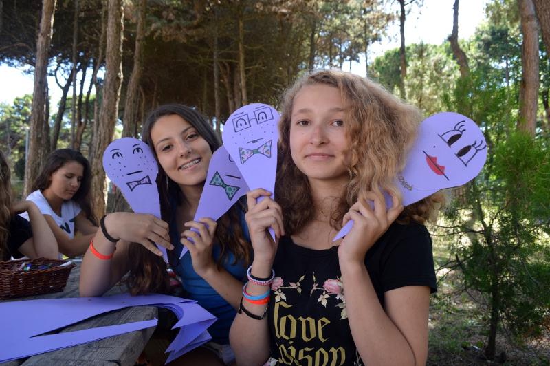 Duas jovens a sorrir enquanto seguram máscaras coloridas com caras desenhadas, participando em atividades de artesanato durante o curso italiano.