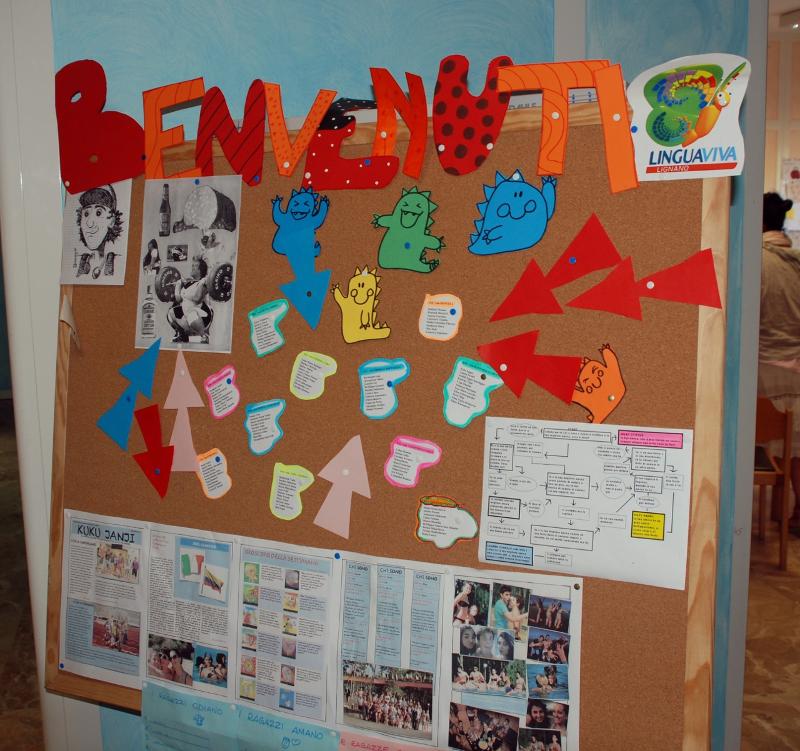 Quadro de avisos colorido com a palavra "BENVENUTI" (bem-vindo) e materiais educativos relacionados com o curso italiano "Lingua Viva".