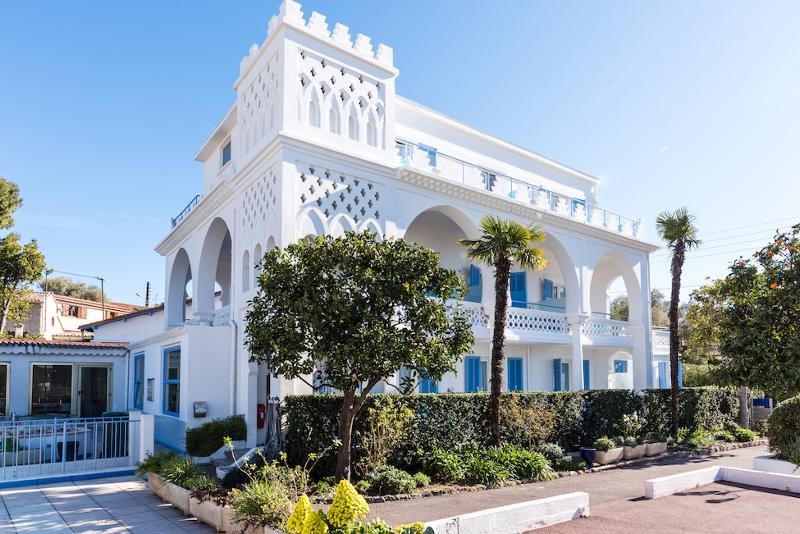 Edifício em estilo mediterrâneo com arcos e detalhes brancos, rodeado por palmeiras e jardim, ideal para um curso de línguas em França