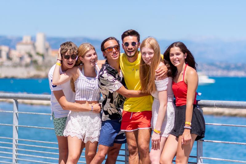 Grupo de jovens sorridentes a posar juntos em frente ao mar, aproveitando o dia ensolarado durante um curso de língua francesa