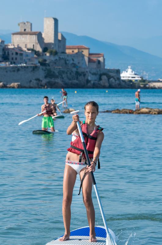 Jovem a praticar paddleboarding no mar, com um castelo histórico ao fundo, durante um curso de língua francesa