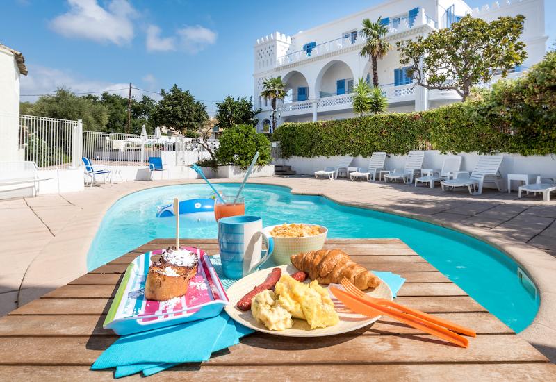 Mesa com um delicioso pequeno-almoço à beira da piscina, com vista para um edifício mediterrânico rodeado de palmeiras