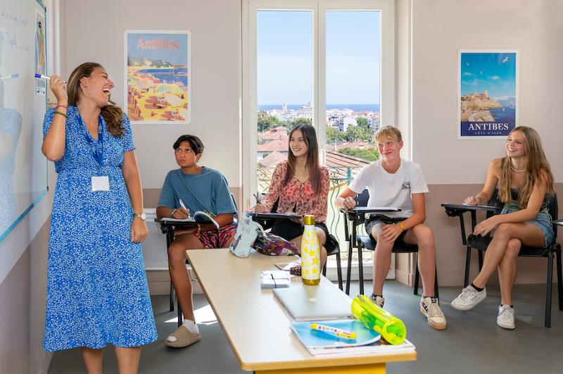 Professora entusiástica a ensinar numa sala de aula, com vista para o mar, enquanto os alunos prestam atenção