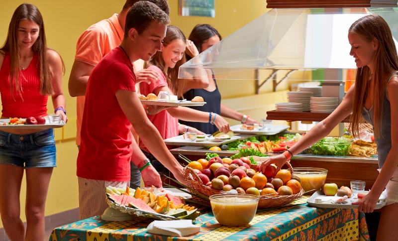 Grupo de jovens a servir-se em um buffet com pratos variados de frutas, saladas e pratos quentes, criando um ambiente de refeição saudável e descontraído durante um curso de língua francesa