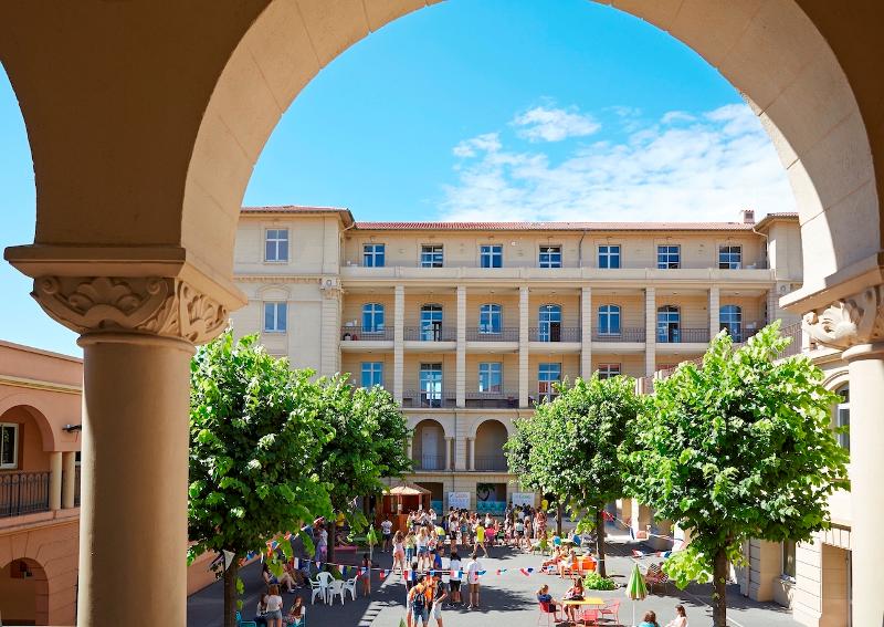 Vista sobre um pátio com muitos jovens a socializar e a participar em atividades, rodeado por árvores e edifícios, num ambiente acolhedor durante um curso de língua francesa