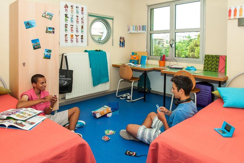 Dois jovens sentados no chão de um quarto colorido, um a jogar às cartas, com um ambiente descontraído e decorado com fotos nas paredes durante um curso de línguas.