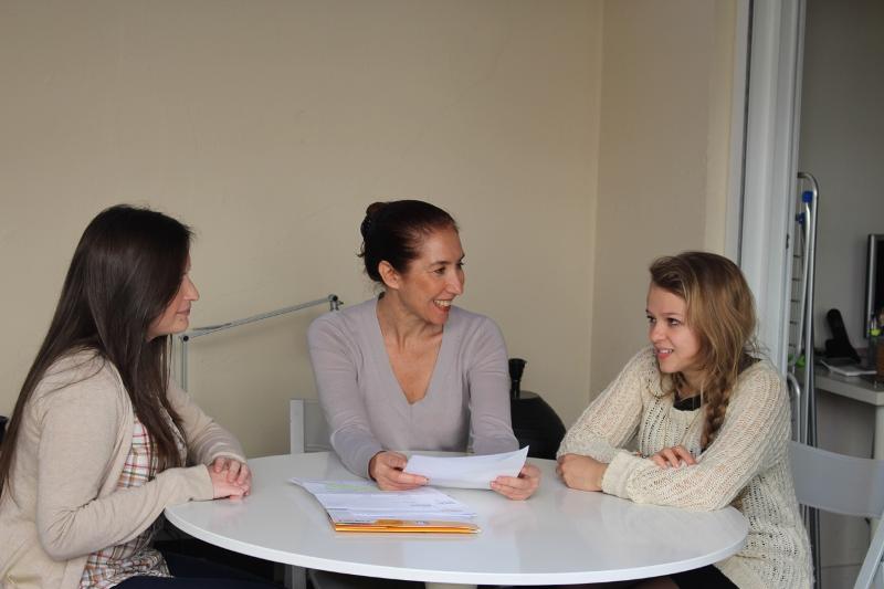 Duas alunas conversam com uma professora ao redor de uma mesa enquanto estudam documentos num curso de espanhol.