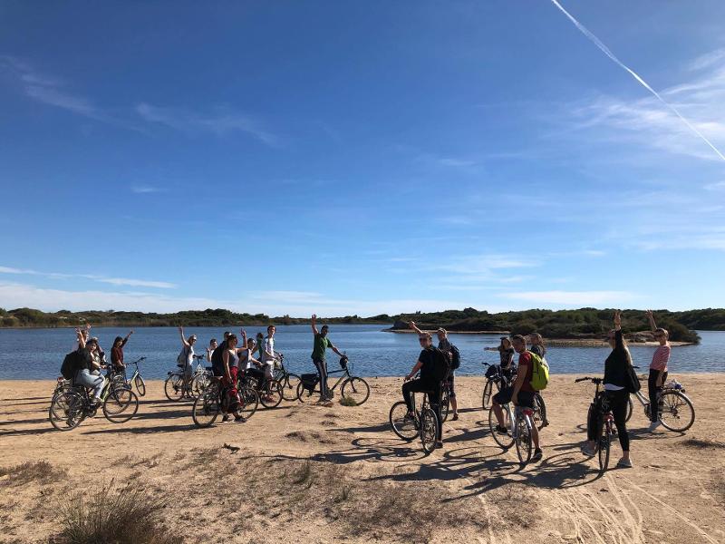 Grupo de pessoas em bicicletas levanta as mãos em celebração junto a uma paisagem de lago, numa atividade ao ar livre de um curso de espanhol.