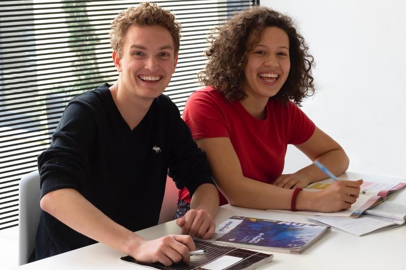 Dois estudantes sorriem enquanto estudam materiais didáticos e utilizam um computador durante um curso de espanhol.