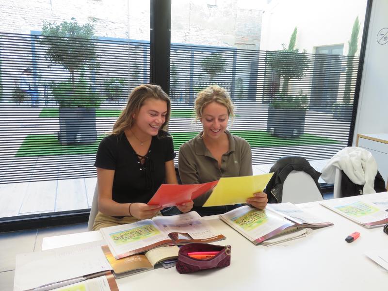 Duas alunas sorriem enquanto leem documentos coloridos durante uma aula de um curso de espanhol.