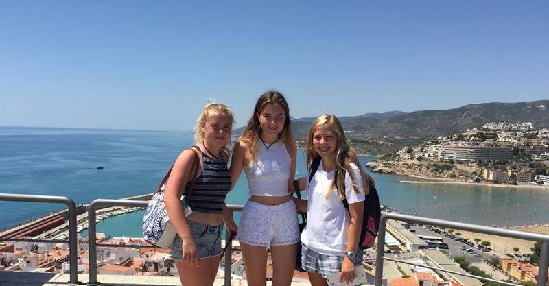 Três jovens posam sorridentes em um miradouro com vista para o mar durante uma excursão de um curso de espanhol.