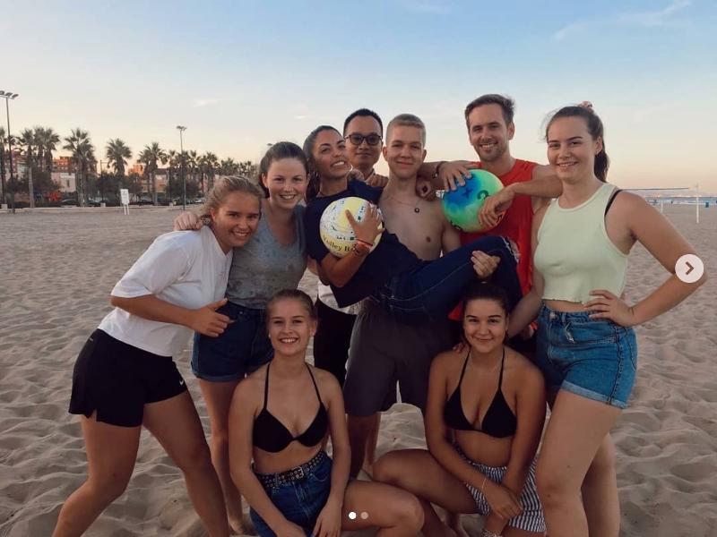 Grupo de jovens na praia posa animadamente com bolas, participando numa atividade recreativa de um curso de espanhol.
