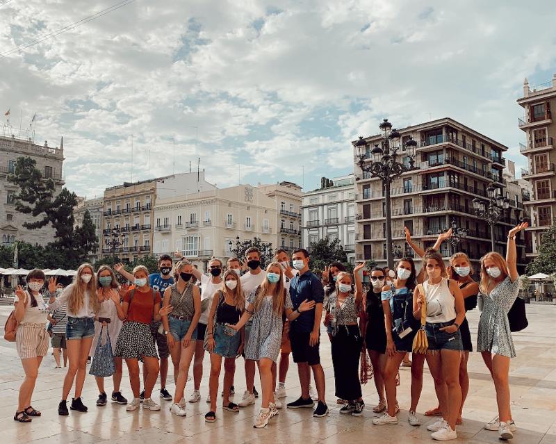 Grupo de estudantes com máscaras faciais posa numa praça central durante uma excursão cultural de um curso de espanhol.